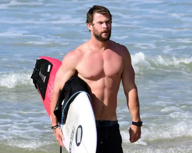 A person at the beach holding a surfboard and walking in the shallow water, wearing black swimming trunks.