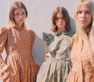 Three women wearing vintage, floral-patterned dresses stand against a white wall, staring at the camera with serious expressions.