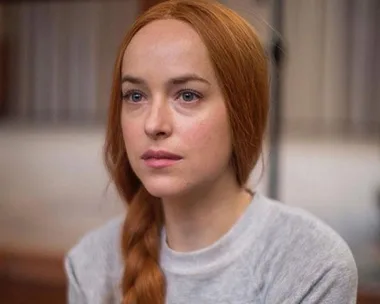 A woman with long red hair in a braid, wearing a light gray top, looking intently ahead.