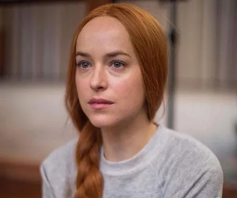 A woman with long red hair in a braid, wearing a light gray top, looking intently ahead.