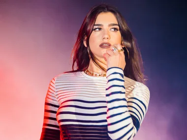 A person with long dark hair in a striped top performing on stage, holding a microphone with colorful lighting in the background.