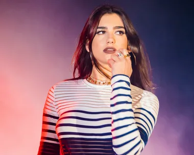 A person with long dark hair in a striped top performing on stage, holding a microphone with colorful lighting in the background.