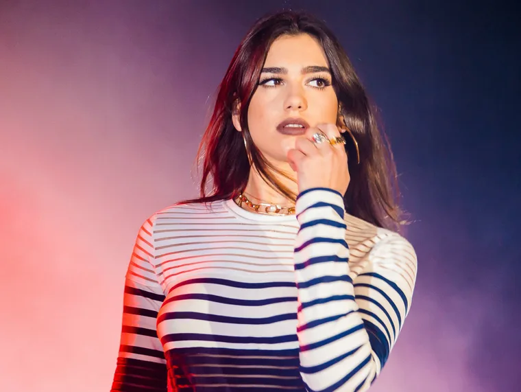 A person with long dark hair in a striped top performing on stage, holding a microphone with colorful lighting in the background.