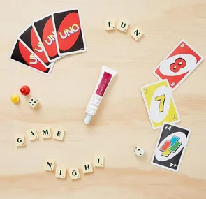 Elucent eye cream surrounded by Uno cards, Scrabble tiles spelling "FUN GAME NIGHT," dice, and game pieces on a table.