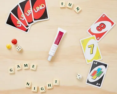 Elucent eye cream surrounded by Uno cards, Scrabble tiles spelling "FUN GAME NIGHT," dice, and game pieces on a table.