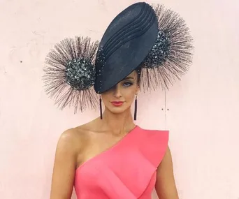 Woman in a pink one-shoulder dress wears an elaborate black fascinator with textured circular elements.