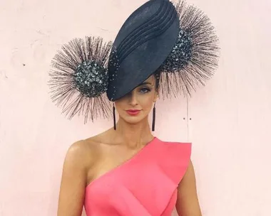 Woman in a pink one-shoulder dress wears an elaborate black fascinator with textured circular elements.