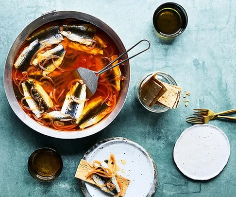 Bowl of sardines in marinade with onions and serving utensils on a blue surface, accompanied by glass jars and plates with crackers.