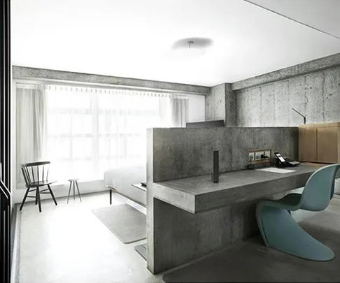 Modern hotel room with minimalist design, featuring concrete walls, large window, bed, desk, chair, and simple decor items.