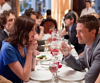 A man and woman gaze at each other holding drinks in a busy restaurant, engaged in a conversation.