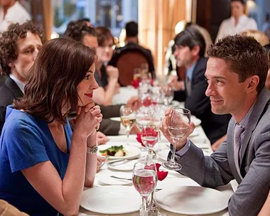 A man and woman gaze at each other holding drinks in a busy restaurant, engaged in a conversation.