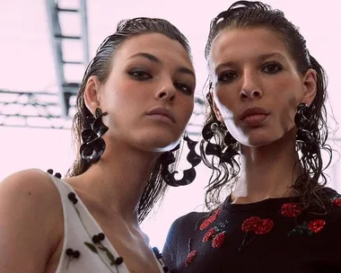 Two models with slicked-back wet-look hair, wearing large black earrings, pose together against a bright background.