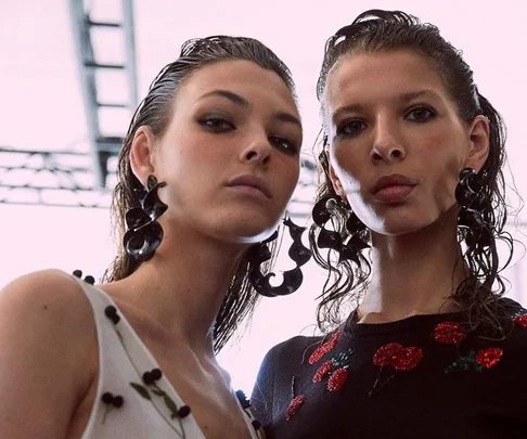 Two models with slicked-back wet-look hair, wearing large black earrings, pose together against a bright background.