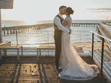 Bride and groom kiss on a sunlit pier with the ocean in the background.