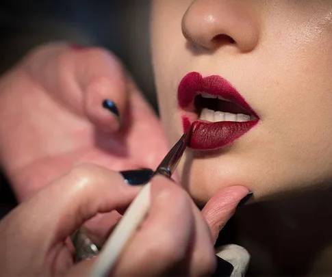 A makeup artist applies dark red lipstick to a woman's lips with a brush.