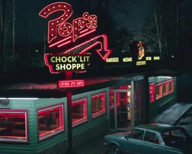 Night view of Pop's Chock'lit Shoppe with neon lights and a vintage car parked outside.