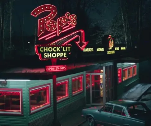 Night view of Pop's Chock'lit Shoppe with neon lights and a vintage car parked outside.