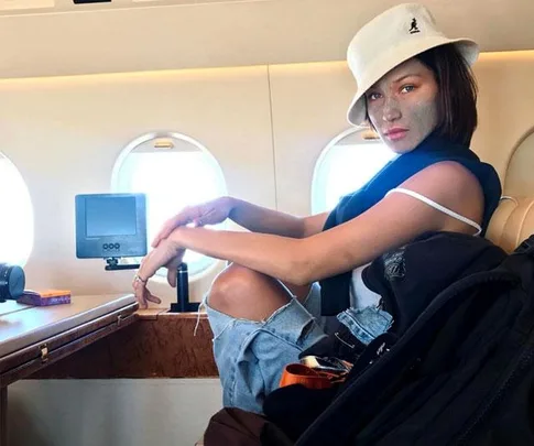 A woman with a face mask sitting in an airplane, wearing a white hat and casual clothes.