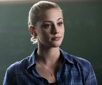 Young woman with blonde hair tied back, wearing a blue plaid shirt, stands in front of a chalkboard, looking thoughtful.