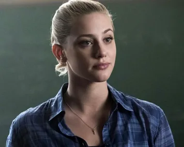 Young woman with blonde hair tied back, wearing a blue plaid shirt, stands in front of a chalkboard, looking thoughtful.