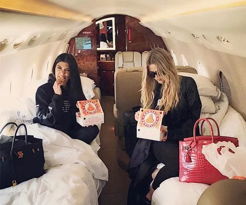 Two women eating Popeyes chicken on a private jet, seated with large handbags and comfortable blankets.