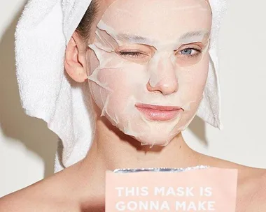 Person winking with a sheet mask on, holding a skincare product that says "This Mask Is Gonna Make."