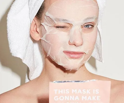 Person winking with a sheet mask on, holding a skincare product that says "This Mask Is Gonna Make."