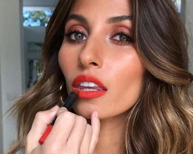 Woman applying vibrant red lipstick, with focus on makeup detailing and soft waves in her hair.