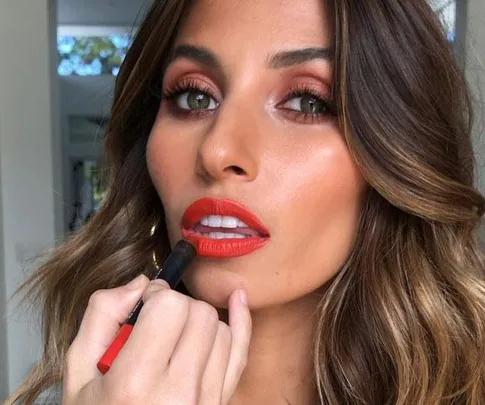 Woman applying vibrant red lipstick, with focus on makeup detailing and soft waves in her hair.