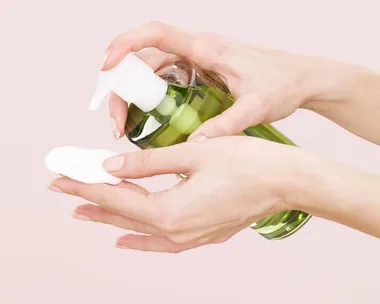 A person dispensing micellar water from a green bottle onto a cotton pad.