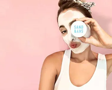 A woman with a white face mask holding a container labeled "SAND & SKY" against a pink background.