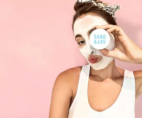 A woman with a white face mask holding a container labeled "SAND & SKY" against a pink background.