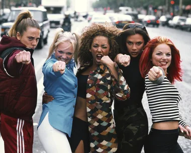 Five women dressed in colorful outfits and posing with their fists out, resembling a famous pop group, standing on a street.