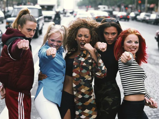 Five women dressed in colorful outfits and posing with their fists out, resembling a famous pop group, standing on a street.