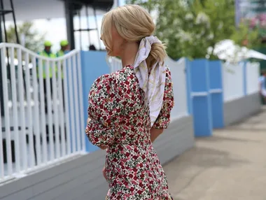 Woman in a floral dress and scarf in her hair, standing outdoors near a fence at an event.