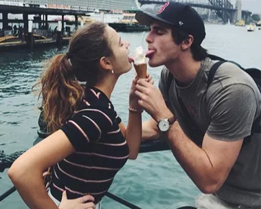 A young couple playfully licking an ice cream cone together by a waterfront, with a bridge in the background.