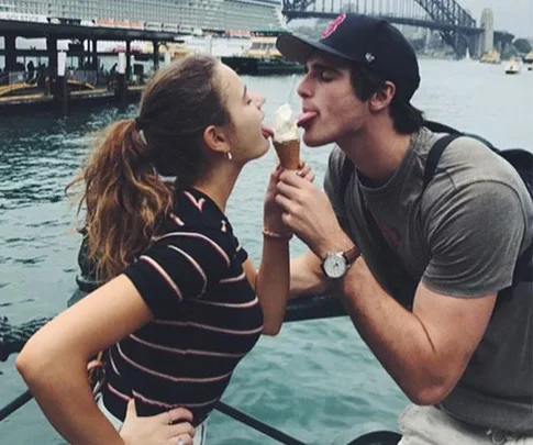 A young couple playfully licking an ice cream cone together by a waterfront, with a bridge in the background.