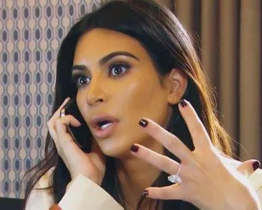 A surprised woman with dark hair holding a phone and gesturing with her hand, showing a large ring.