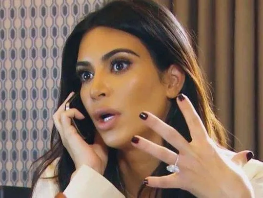 A surprised woman with dark hair holding a phone and gesturing with her hand, showing a large ring.