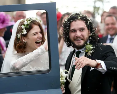 Bride and groom, dressed elegantly, smile and celebrate with guests outside a car on their wedding day.