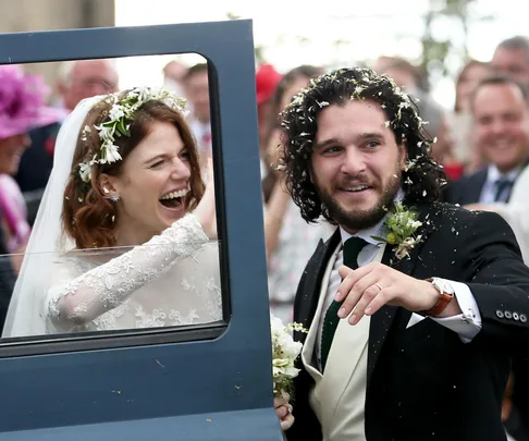 Bride and groom, dressed elegantly, smile and celebrate with guests outside a car on their wedding day.