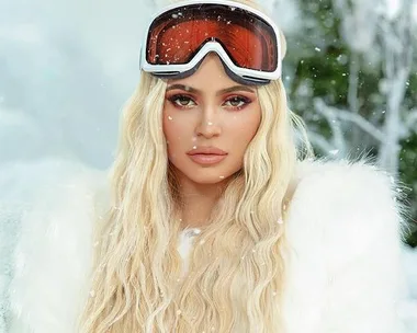 Woman with long blonde hair in a snowy landscape wearing a white fur coat and ski goggles.