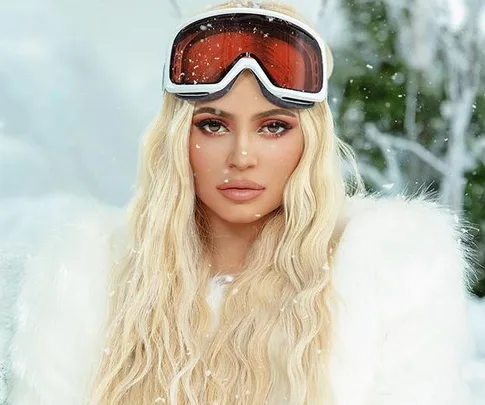 Woman with long blonde hair in a snowy landscape wearing a white fur coat and ski goggles.