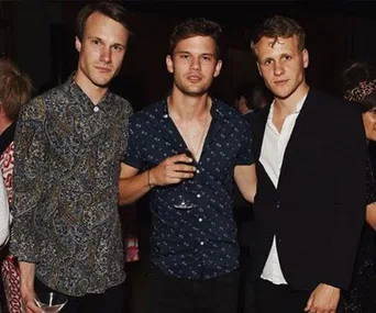 Three young men from "Mamma Mia! Here We Go Again" standing together at a party, one holding a drink, dressed in casual and formal attire.