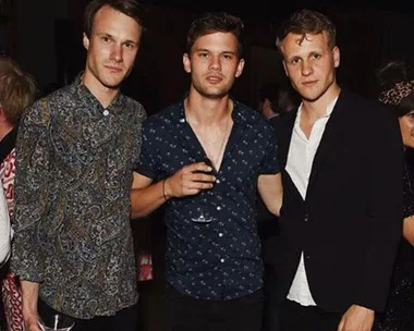 Three young men from "Mamma Mia! Here We Go Again" standing together at a party, one holding a drink, dressed in casual and formal attire.