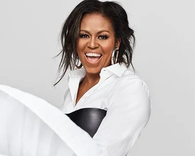 Michelle Obama smiling, wearing a white blouse and large hoop earrings against a plain background.