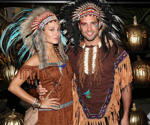 Two people in Native American-themed costumes with feathered headdresses and fringed outfits at a Halloween event.