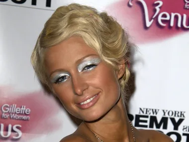 Person with blonde hair, blue eyeshadow, and glossy lips smiles at a Gillette Venus event backdrop.