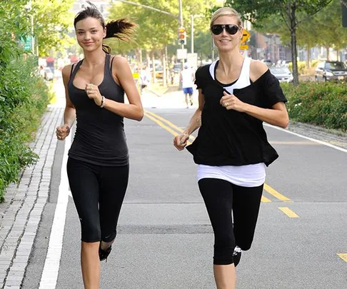 Two women jogging on a tree-lined urban road, wearing athletic clothing and smiling.