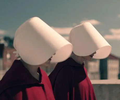 Two women from "The Handmaid's Tale" wearing red cloaks and white bonnets, walking outdoors in daylight.